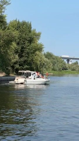 A white motorboat with a canopy and several passengers is moving on a river. The boat has people on deck, some of whom appear to be wearing life jackets. The riverbank to the left is lined with green trees. In the background, a concrete bridge spans the river, with cars and trucks visible on it. The sky is clear and blue. The water surface is rippled.