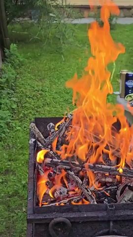 The video shows a close-up of a barbecue grill filled with burning wood. Flames are actively burning and flickering, with visible embers and ash at the bottom of the grill. The grill itself is made of dark metal and appears to be outdoors, with green grass and foliage visible in the background. The overall scene suggests a casual outdoor cooking setting.