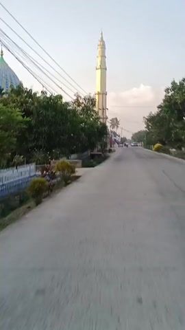 The video shows a tall, slender minaret, likely part of a mosque, standing prominently against a pale sky. The minaret has multiple tiers and a decorative finial at its apex. It is a light yellow or cream color with some reddish-brown accents on the lower sections. Trees with green foliage line the sides of a paved road that curves gently into the distance. Several utility poles with a dense network of wires are visible, some running in front of the minaret and the mosque. A building with a dome, presumably the mosque itself, is partially visible to the left of the minaret. Small structures, possibly shops or stalls, are situated along the roadside near the base of the minaret. A few vehicles are seen on the road in the background, and a person or two are visible near the roadside structures. The lighting suggests it is either early morning or late afternoon.
