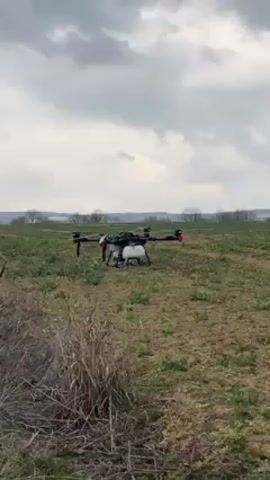 The video shows a drone lifting off from a grassy field. The drone is black and white with red accents and has a tank attached underneath. The drone takes off slowly and hovers in the air. The background includes a grassy field, trees, and a cloudy sky.