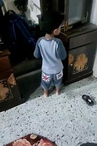 A young child wearing a blue and gray long-sleeved shirt and denim shorts with British flag patches on the back pockets is standing in front of a dark wooden cabinet. The child is wearing a black cap and facing away from the camera, looking into the open cabinet. The cabinet has an ornate design on its doors and a mirror on the right side. A blue backpack is partially visible on the left side of the cabinet, and a black sandal lies on the floor to the right of the cabinet. The floor appears to be tiled with a speckled pattern. The child's reflection is visible in the mirror of the cabinet. The child's left hand is resting on the edge of the cabinet, and their right hand is reaching into the cabinet. A small blue toy truck is visible inside the cabinet. The lighting in the room is dim.