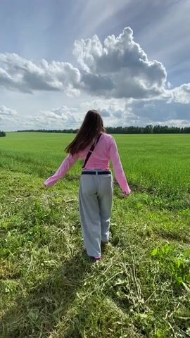 A young woman with long brown hair is running in a grassy field under a cloudy sky. She is wearing a pink long-sleeved shirt, grey pants, and sunglasses. The field is green and lush, with a line of trees in the distance. The sky is blue with large, white, fluffy clouds. She turns around and smiles at the camera.