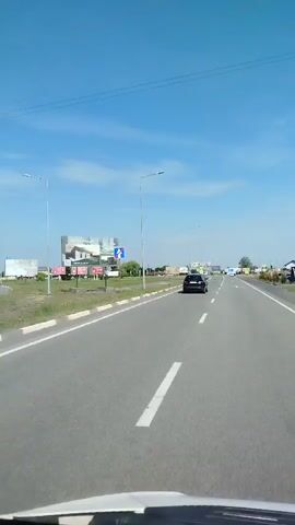 The video shows a view from the dashboard of a car traveling on a highway. The road is paved and marked with white lines. There are several cars on the road, including a dark-colored car in front of the camera. On the sides of the road, there are billboards, streetlights, and some buildings. The sky is blue with some clouds. The video appears to be filmed during the daytime. The camera is mounted inside the car, providing a first-person perspective of the drive. The car is moving forward, and the scenery is passing by. The video shows signs with arrows indicating directions for lanes.