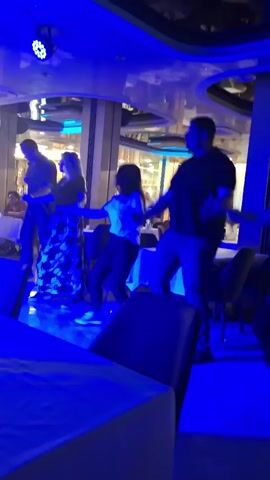 Four adults are line-dancing inside a restaurant or entertainment venue under color-changing lights. They hold hands, stepping side to side in unison. The interior features tables with white tablecloths and upholstered chairs. The lighting alternates between blue, magenta, red, and green. There are other people in the background, seated at tables.