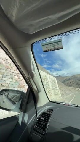 The video is filmed from inside a vehicle looking out through the windshield at a mountainous landscape. The sky is partly cloudy with patches of blue. The road ahead curves to the right and appears to be paved. The mountains are steep and rocky, with sparse vegetation. There is a construction site visible on the side of the road with yellow heavy machinery. A sticker is visible on the upper part of the windshield, which appears to be an emergency road assistance sticker with text and a phone number.