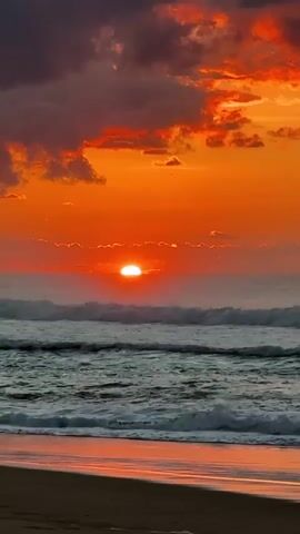 A vibrant sunset over the ocean. The sun, a bright orange orb, is low on the horizon, casting a warm glow across the sky. Wispy clouds are tinged with shades of orange, pink, and purple. The sea is choppy, with waves rolling towards the shore. The wet sand in the foreground reflects the colors of the sky. The video is shot from a beach perspective, looking out towards the ocean and the setting sun.