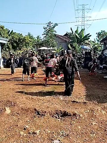 The video shows a traditional dance performance taking place outdoors on a dirt surface. Several people are performing, dressed in traditional Indonesian clothing. Some are in black outfits while others have more colorful attire. One dancer is lying on the ground. The performance appears to be taking place in a rural area, with houses and trees in the background, and an audience of onlookers. A large electrical tower is visible in the background. The sky is clear and blue, suggesting it is a bright, sunny day. There is a sound system nearby, and the performance seems to be accompanied by music.
