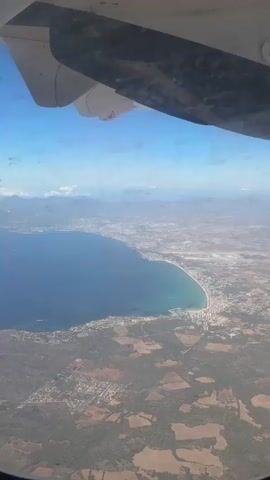 This is a first-person perspective video taken from an airplane window. The viewer sees a body of water, possibly the ocean or a large lake, to the left. The water is a deep blue and meets a sandy coastline with buildings along it. To the right of the water is land with a mix of agricultural fields, green areas, and some buildings. In the distance, there are mountains or hills. The top of the airplane's wing is visible in the foreground, obscuring a portion of the view. The sky is blue with some wispy clouds. The video is shaky, suggesting movement. There are no people or specific actions visible beyond the general view of the landscape from the aircraft.