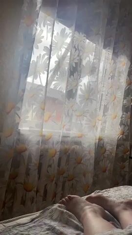 A person is lying in bed in a room filled with light. The person's bare feet are visible in the foreground. The bed has a flowered sheet. Behind the bed, there is a window covered with a daisy-patterned curtain, allowing sunlight to stream into the room. The window faces a building with a pinkish-red roof.