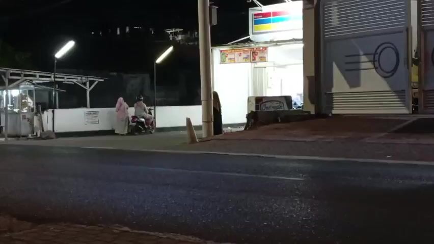 The video depicts a nighttime scene on a road adjacent to a small convenience store. A few people are visible near the store entrance, with two individuals on a motorcycle. The road is wet, reflecting the streetlights. Several vehicles, including motorcycles and a van, are captured in motion, creating motion blur. The convenience store has bright interior lighting and signage, including the familiar colors of a large convenience store chain. The overall impression is that of a typical urban street scene at night, with ongoing traffic and commercial activity.