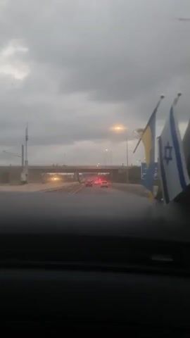 The video shows a view from inside a vehicle, looking forward through the windshield. The sky is overcast and gray. In the distance, a highway overpass is visible, with traffic moving beneath it. Several cars are on the road ahead, and one appears to be a police car with flashing blue and red lights. Two flags are mounted on the right side of the vehicle's dashboard, partially obscuring the view. One flag is blue and yellow, and the other is the Israeli flag. The overall impression is of a drive on a cloudy day, possibly in Israel, with a police presence on the road.