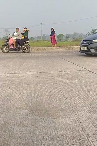 The video shows a grey car parked on the side of a road. A person wearing a red and pink sari stands near the car. The car's trunk is open. In the background, there are fields and trees, and a motorcycle is seen passing by. The road is made of concrete with visible lines. The sky is overcast.