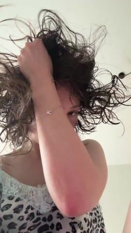 A woman with curly, wet, dark hair stands in a bathroom. She is wearing a white lace-trimmed camisole with a black and white animal print. She is looking directly at the camera with a serious expression and is running her fingers through her hair, tousling it. The background is a white wall, and a portion of a bathroom light fixture can be seen.