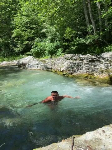 A man is standing in a shallow body of water that appears to be a river or creek. He is shirtless and wearing shorts. His arms are outstretched to his sides, and he is looking towards the camera. The water is clear and has a greenish-blue tint. The surrounding environment is a wooded area with lush green trees and rocky terrain. The lighting suggests it is daytime, with sunlight filtering through the trees.