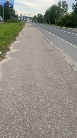 A first-person perspective shot shows a person walking on a cracked concrete sidewalk next to a two-lane road with cars driving on it. The sidewalk is bordered by a strip of green grass on the left. On the right, the road is lined with trees and some distant buildings. A billboard with a picture of a person in uniform is visible on the far left. The sky is overcast.