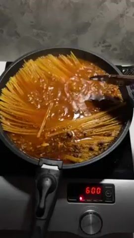 A top-down, medium shot shows a pan of spaghetti cooking on a stovetop. The spaghetti is yellow and submerged in a red, bubbling sauce containing ground meat. The stovetop is black and has a digital display showing "600". A person’s hand appears, holding a wooden spoon, and pushes the spaghetti down into the sauce. The background is a gray wall.