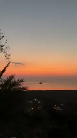 The video presents a scenic vista at dusk. Above the treeline, the sky transitions from pale grey at the top to a fiery orange as it meets the horizon. The ocean reflects the sunset's hues, with a small, silhouetted island breaking the horizon line. In the foreground, the dark silhouettes of trees and foliage, notably a palm tree on the left, frame the view. The lights of a settlement are visible in the valley below the horizon. Human voices can be heard in the background.