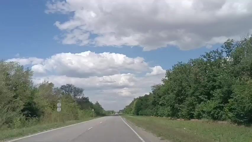 The video shows a dash cam view of driving on a two-lane asphalt road surrounded by lush green trees and vegetation on both sides. The sky is blue with scattered white clouds. There are a few cars on the road, including one red SUV driving in the opposite direction and another dark car further ahead. The road appears to be in a rural or suburban area.