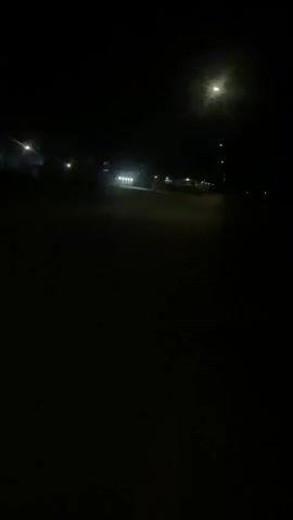 The video shows a dark, outdoor scene at night. The camera appears to be moving forward along a paved path or road. On the left side of the path, there are several bright lights illuminating buildings or structures. A person on a scooter or similar vehicle is visible moving away from the camera in the distance. There are also what appear to be large white rocks or planters along the left side of the path. The right side of the path is bordered by grass or vegetation. A street light is visible in the upper right portion of the frame, casting a bright glare. The overall environment suggests a residential area, park, or campus setting at night.