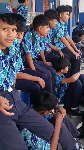 The video shows a group of male students sitting on a ledge and on the ground outside a building. The students are all wearing similar uniforms: blue shirts with a white and green pattern and dark blue pants. Many of them are wearing backpacks. The building appears to be a school, with blue and white trim. The students are mostly looking in different directions, with some talking and others appearing to be looking at something out of frame. One student in the foreground is smiling and laughing. Another student in the foreground is leaning down and appears to be looking at their hands. In the background, a white car is visible driving down a street. The ground is paved with concrete slabs.