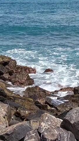 The video shows waves crashing against a rocky shoreline. The water is a clear blue-green, and the rocks are dark and rough. The waves are white and foamy as they break on the rocks. The sky is a clear blue.