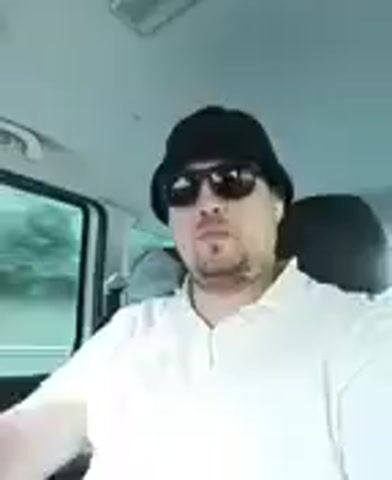 This media file captures a close-up, selfie-style shot of a man from the chest up, seated inside a vehicle. The man is wearing a black bucket hat and dark sunglasses, obscuring much of his face. He has a light goatee and appears to be wearing a light-colored polo shirt. A glimpse of white earbuds can be seen in his right ear.

The setting is clearly the interior of a car, with a headrest visible behind the man's right shoulder and a window to his left, through which blurry green foliage can be seen, suggesting movement through a rural or wooded area. The sky appears overcast, and the lighting suggests it is daytime, possibly late afternoon or a heavily clouded day, contributing to a slightly muted ambiance.

The man's gaze is directed slightly off-camera, and his expression is neutral. The overall impression is that he is either driving or a passenger in a vehicle, perhaps recording a brief video update or simply capturing a moment during his journey. No text is visible in the media file. The context of Turkovice, Czechia, places this scene within that specific geographical location.