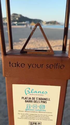 This video features a close-up of a brown metal post on a sandy beach. The post has engraved text that reads "take your selfie" and a small triangle shape at the top. Below the text, there is a white label with the word "Blanes" in a stylized blue font, followed by "WELCOME TO THE COSTA BRAVA!". Further text on the label indicates "PLATJA DE S'ABANELL BARRI DELS PINS". There are also social media icons for Instagram and Facebook, along with a QR code and a website address.

In the background, a beach stretches out with gentle waves lapping at the shore. The sky is clear and blue, suggesting good weather. The lighting indicates it is likely late afternoon or early evening, with the warm glow of the sun casting long shadows. A few people can be seen in the distance walking along the beach and near some buildings on a hill overlooking the coastline. The overall scene evokes a relaxed and tourist-friendly atmosphere. The object in the foreground is likely a photo opportunity or a marker for a specific scenic spot on the beach.