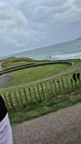 The video shows a scenic view of a coastal area on an overcast day. In the foreground, there is a gravel path. Beyond the path, a wooden fence runs across the frame, with what appear to be two pieces of dark clothing hanging from it. Behind the fence is a circular grassy area with a low stone wall bordering it. This circular area appears to be some kind of amphitheater or gathering space. Further in the background, the ocean is visible with moderate waves and whitecaps. Two yellow warning signs are present near the edge of the grass overlooking the ocean. The sky is cloudy and gray, and a distant headland can be seen on the right side of the frame. The overall atmosphere is one of natural beauty, perhaps a bit wild and windswept.