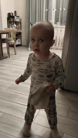 A toddler walks through a room holding a stuffed animal. The toddler is wearing pajamas and slippers. The room is decorated with furniture and a window with curtains. The video is shot from a medium angle, and the lighting is dim.