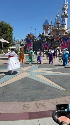 This media file captures a lively outdoor scene in front of Sleeping Beauty Castle at Disneyland. The sun is shining brightly, indicating a clear, warm day, likely midday. A group of performers are engaged in a musical performance.

On the left, a woman dressed as Mary Poppins, complete with a white hat and dress and holding a pink umbrella, twirls. To her right, a brass band is assembled. The band members are dressed in colorful, coordinated outfits: one in bright yellow, another in orange, one in green, and another in purple, playing various brass instruments like a tuba and a saxophone. Further to the right, other performers in matching colorful costumes are visible, suggesting a larger ensemble.

The backdrop is dominated by the iconic pink and blue turrets of Sleeping Beauty Castle, adorned with decorative banners. The ground features a large compass rose design, with star accents. In the background, other park guests can be seen walking and observing the performance.

The overall atmosphere appears festive and joyful, with the performers radiating energy and the setting exuding classic Disney magic. The visible text on the ground appears to be part of a circular inscription, potentially reading "DREAMS" or similar words related to the park's theme.