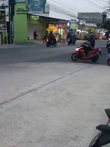 The video shows a street scene with several motorcycles and one car. The street is paved, and there are buildings on the right side of the road. The buildings appear to be shops or businesses, with signs and awnings. The sky is overcast, suggesting it might be evening or an overcast day. Several people are riding on the motorcycles. One motorcycle in the foreground is red and is being ridden by a person wearing a helmet. Another motorcycle in the background is white and is also being ridden by a person wearing a helmet. A silver car is also visible, driving on the street. The overall atmosphere is that of a typical urban or suburban street with traffic.