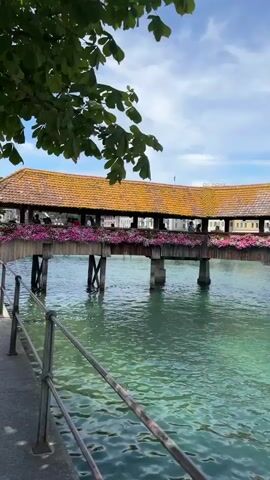 A video shows a covered wooden bridge decorated with pink flowers spanning a body of clear, turquoise water. People are walking on the bridge and along a paved walkway beside the water. The bridge has a reddish-brown tiled roof and wooden supports. Green leaves from a tree overhang the top left of the frame. In the background, a building with a red-domed roof is visible against a blue sky with scattered white clouds. The water is calm with gentle ripples. A metal railing runs along the edge of the walkway.