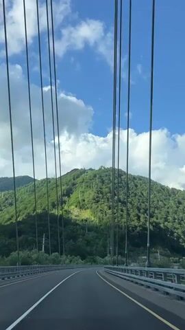 This is a POV shot from a vehicle driving on a suspension bridge. The bridge has multiple lanes and guardrails on the side. Vertical cables of the suspension structure are visible, some of which are illuminated by streetlights. Beyond the bridge, a lush green mountain fills the background. The sky is blue with scattered white clouds. The time stamp is 00:00 to 00:05.