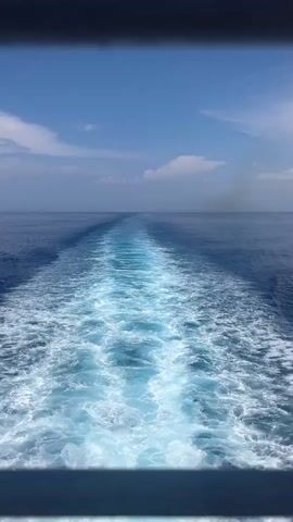 This is a vertical video shot from the stern of a moving vessel on a clear, sunny day. The camera is looking out over the water at the turbulent wake left by the vessel. The wake is a frothy white trail that gradually fades into the deep blue ocean. The sky is a vibrant blue with a few scattered white clouds. The horizon is visible in the distance, where the sea meets the sky.