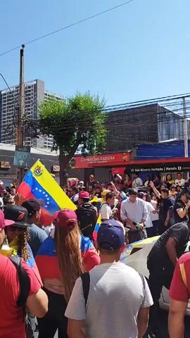 A crowd of people march down a city street, holding and wearing Venezuelan flags. The sky is clear and the sun is shining. The crowd is dense, with a mix of men and women, most wearing casual clothing. Some are wearing baseball caps. The flags are the main visual element, adding bursts of yellow, blue, and red to the scene. A band is playing brass instruments, and a person seems to be giving an interview using a microphone. The crowd moves along the street and sidewalks, surrounded by buildings, power lines, and other urban elements.  A sign indicates that there is a subway station nearby.