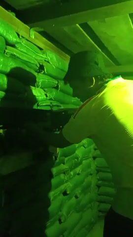 A person wearing a hat and a light-colored shirt is aiming a weapon. The person is positioned behind sandbags stacked to form a wall. The environment is illuminated with green lighting. The video shows the person firing the weapon multiple times.