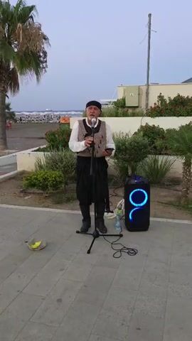 A man in traditional clothing is singing into a microphone with a portable speaker next to him. The man is wearing a white shirt, a vest, black pants, and a black headscarf. He is standing on a paved walkway with some greenery and palm trees behind him. In the background, there is a beach with umbrellas and people. The sky is overcast.