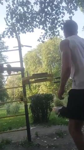A person is performing dips on a parallel bar exercise station outdoors. The person is wearing a white tank top and black shorts. The exercise station is made of yellow metal and is situated in a grassy area with trees in the background. The sky is visible through the leaves of a tree in the foreground.