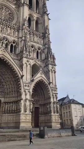 The video shows a low-angle, wide shot of the exterior of a large, ornate Gothic cathedral. The facade is elaborately decorated with sculptures, arches, and windows. In the foreground, a paved square with cobblestone patterns is visible. A few people are walking across the square. One person, a young man in a blue jacket and dark pants, is the most prominent figure, walking towards the cathedral. The sky above is overcast. To the right of the cathedral, a portion of a modern building is visible. The camera seems to be slightly tilted upwards, emphasizing the grandeur of the cathedral.