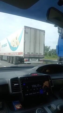 This is a first-person perspective video from inside a vehicle. The driver is looking at the dashboard display which shows a radio interface with the frequency 106.10. Outside the car, a traffic jam is visible on a highway. Several large trucks and a bus are in front of the vehicle. One truck on the right side of the frame has an advertisement on its side featuring a cartoonish drawing of potatoes in a hammock. Another truck on the left side of the frame has text that appears to be in Russian, possibly indicating "OOO VELES". Further ahead, there is a large construction vehicle, possibly an excavator, being transported on a flatbed truck, and a white bus. The sky is overcast.