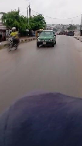 The video shows a first-person perspective of someone riding a motorcycle on a wet, paved road. The sky is overcast and it appears to be raining lightly, with water visible on the road surface and spray from the motorcycle's tires. On the left side of the road, there are small buildings that appear to be shops or stalls, with some trees and utility poles visible. Several other motorcycles and a green vehicle are also on the road, moving in the same direction. The overall environment suggests a rural or suburban area during daytime.