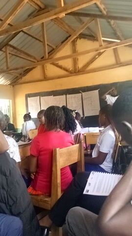 The video shows a group of people, primarily young adults, seated in what appears to be a classroom or training room in Mwingi, Kenya. They are seated on wooden chairs at wooden desks, facing a front wall, though the camera perspective is from within the group, looking towards the front and slightly to the side.

Several individuals are visible, some looking down at notebooks or papers on their desks, suggesting they are engaged in learning or taking notes. One person in the foreground on the right side of the frame is holding a notebook with visible text, although the content is not decipherable. Another individual is speaking, and the audio includes the phrase "how will I."

The room has a rustic appearance with a visible wooden beam structure supporting a roof that appears to be made of corrugated metal sheets, typical of constructions in the region. Papers are tacked to the wall at the front of the room, possibly charts or lesson materials, but the details are unclear. Natural light illuminates the room, indicating it is daytime. The overall atmosphere suggests a focused learning or workshop environment.