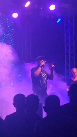 A man wearing a black t-shirt and a black baseball cap is on a stage, holding a microphone. He is singing and gesturing with his right hand. There is smoke on the stage, and purple lights are illuminating the scene. In the background, another person is playing a guitar. The stage is set up for a live performance with lights and speakers. There are silhouettes of people in the foreground, watching the performance.