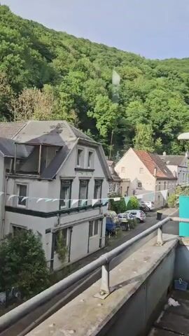 The video presents an elevated view of a residential street in Altena, Germany. In the foreground, a sturdy metal railing suggests the vantage point is a balcony or terrace. The street below is lined with several houses, primarily featuring traditional European architectural styles with gabled roofs. One prominent house in the immediate left foreground has white walls, dark window frames, and decorative elements, while another to its right is a lighter cream color with a reddish-brown tiled roof.

A steep hillside, densely covered with vibrant green trees, forms the prominent backdrop, suggesting a natural and perhaps rural or semi-rural setting. Along the street, several cars are parked, including a light blue van and multiple white or silver sedans. A string of white and light blue pennant banners is stretched across the street, adding a decorative or festive touch. The scene is brightly illuminated, indicating daytime with clear or partly cloudy weather. No people are visible in the frame, and there is no discernible text on any objects or buildings.