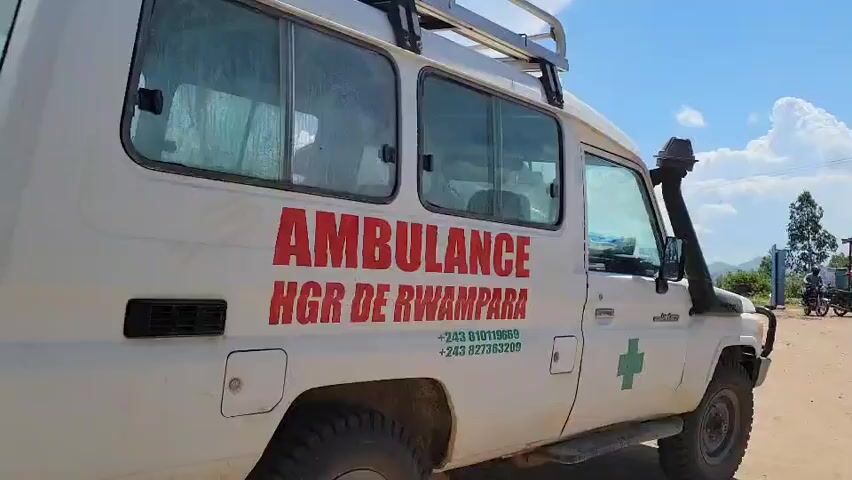 A white ambulance, likely an off-road capable vehicle such as a Toyota Land Cruiser given its high clearance and snorkel, is parked on a dirt road. The vehicle features a roof rack. Large red lettering on its side identifies it as an "AMBULANCE" and specifies "HGR DE RWAMPARA". Below this text, two phone numbers are listed: "+243 810119669" and "+243 827363209". A green cross symbol is visible on the lower right side of the ambulance. Faint silhouettes of individuals can be seen through the front and rear passenger windows. The setting is outdoors under a bright, partly cloudy sky during daylight hours. In the background, there are green trees, lush vegetation, and distant hills or mountains. A motorcycle and other figures are visible further along the dusty road. This scene takes place in Tchere, Democratic Republic of the Congo.