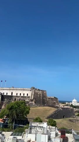 This is a vertical video of a city skyline with a large stone structure in the foreground. The sky is clear and blue. The stone structure appears to be a fort or castle. In the background, there are buildings of various sizes and colors, as well as trees. There are some cars driving on a road. The video pans slightly to the right, showing more of the city.