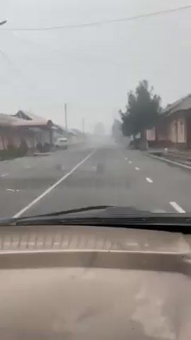 The video shows a first-person perspective from inside a vehicle driving down a straight, paved road. The road has white lines marking the lanes. The environment is hazy or foggy, creating low visibility. On either side of the road are rows of single-story buildings with varying architectural styles. Some appear to be residential houses. Several parked vehicles are visible along the sides of the road. There are also trees sparsely planted along the road. The dashboard of the vehicle is visible in the foreground, indicating the camera is mounted inside the car. The car appears to be moving at a steady pace.