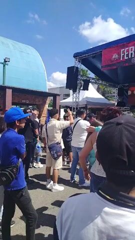 The video shows a crowd of people watching a performance on a stage outdoors at a "Feria Los Altos" event. The stage has a banner with the event name and an Instagram handle. The people are casually dressed and appear to be enjoying the event. Several people are looking at the stage and one person takes a picture with his cell phone. The weather appears to be sunny and clear.