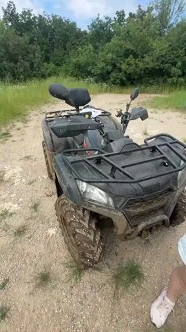 A black all-terrain vehicle with muddy tires is parked on a dirt path. A woman wearing light blue denim shorts and a black top stands to the right, holding a smartphone to her face, seemingly filming. A man in a black t-shirt is briefly visible near the rear of the ATV. The camera then pans to show a wide panoramic view from a hilltop, revealing a vast valley with green fields, forested areas, a distant river, and scattered buildings under a partially cloudy sky.