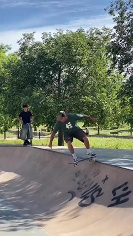 A daytime video shows a man skateboarding in a concrete skate park bowl under a clear blue sky. He wears a dark green t-shirt, dark shorts, a cap, and white sneakers.

At the beginning of the video, the man is standing on the edge of the bowl with his skateboard. Another person, dressed in a black t-shirt and light-colored pants, stands further back on a scooter on the flat ground surrounding the bowl. Lush green trees line the background.

The skateboarder drops into the bowl, smoothly riding down one side and up the opposite wall, where graffiti is visible on the concrete surface, including a colorful, stylized graphic. He then turns and rides back down into the bowl before riding up the initial side again, reaching close to the coping before turning back down. The man on the scooter remains in the background, observing. The concrete bowl has visible graffiti, and a sign partially visible in the background reads "VULCANI."